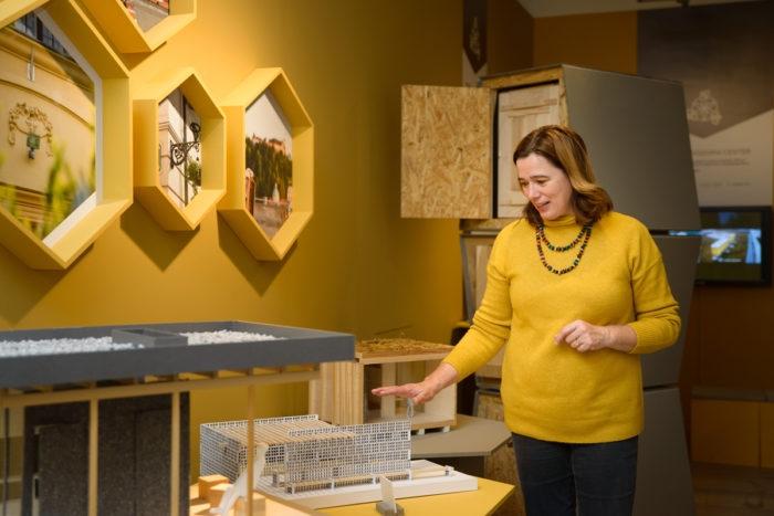 A woman stands over a small model of a beehive. Behind her is a yellow wall with honey-comb shaped pictures. 