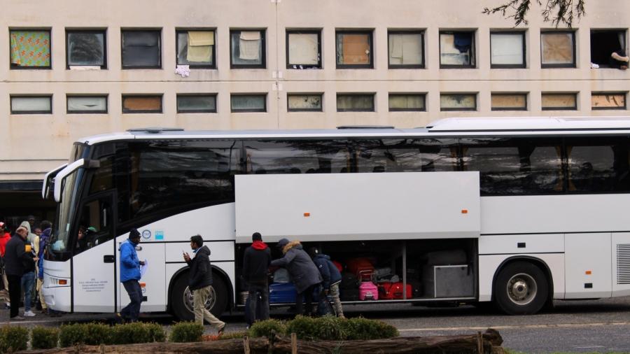People load a white bus with luggage in front of a cement building. 