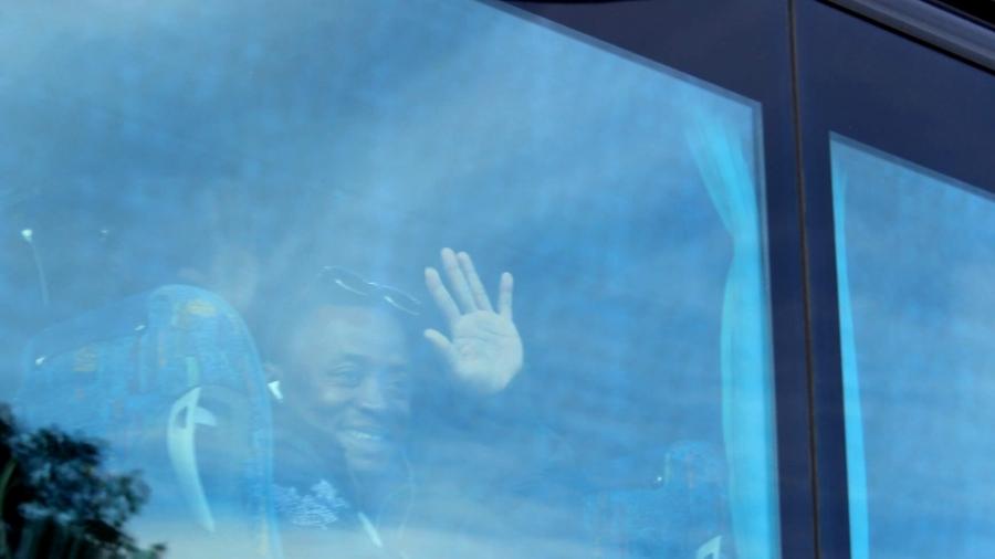 A young African man waves goodbye from the window on a bus. 