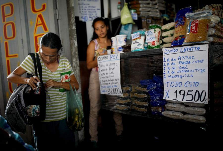 A woman is shown at a market with signs in Spanish for various groceries in front of her.