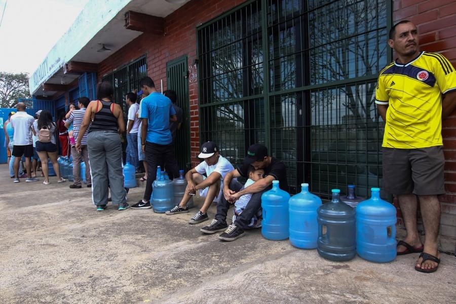 People a showning standing in line next to a brick building waiting with empty jugs.