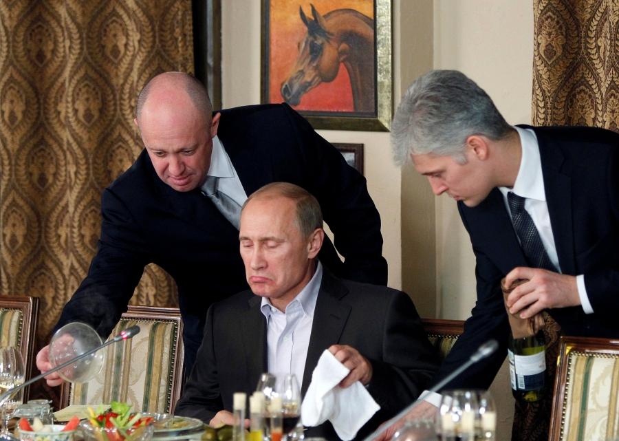 Putin sits at a desk while a man on either side stand behind him 