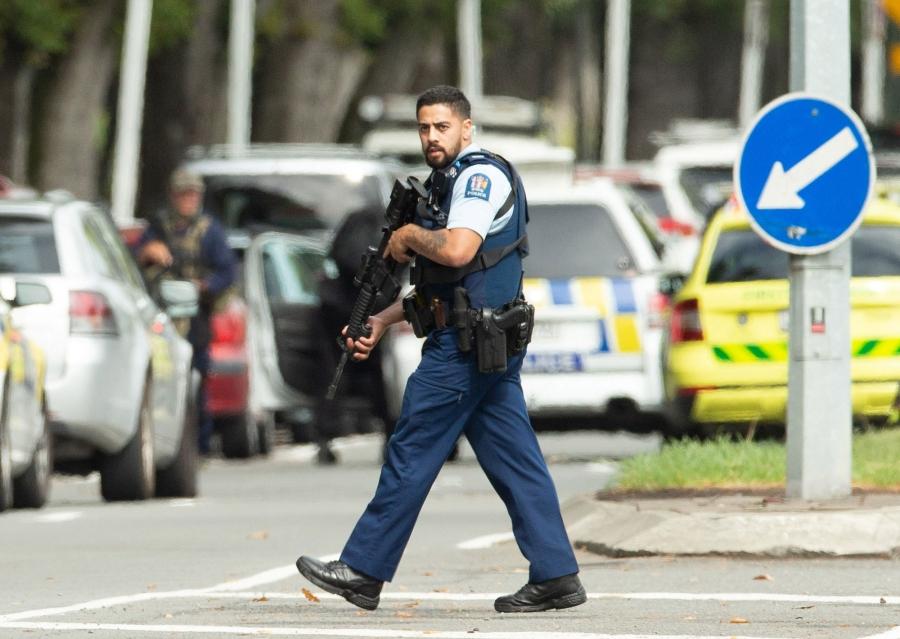 A police, in a short-sleeved uniform and carrying a rifle, is shown walking the street in Christchurch, New Zealand.
