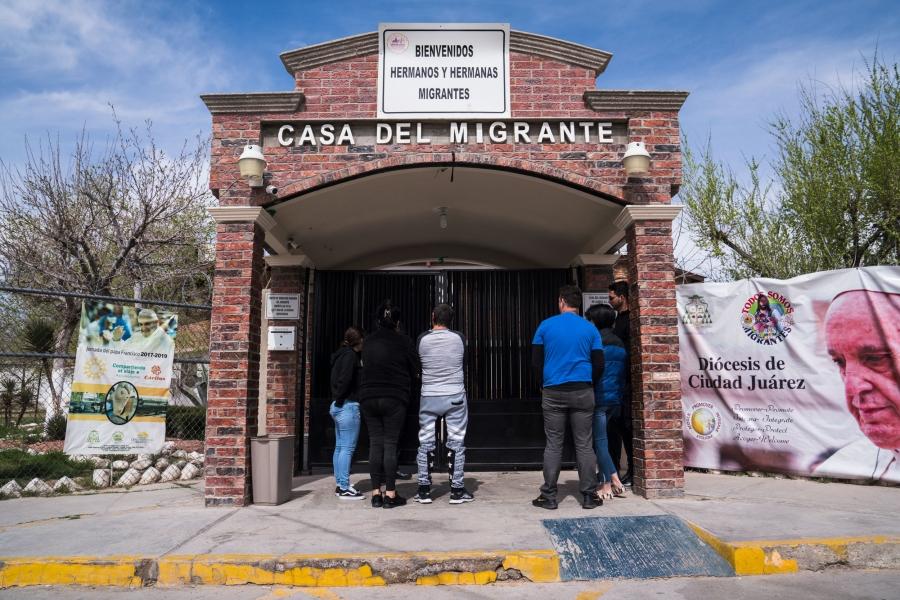 Several migrants from Cuba are shown with their backs to the camera in front of a brick building with a sign that reads "Casa Del Migrante."