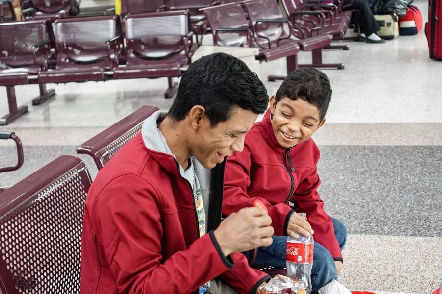 A father and son sit together on a bench in a bus station.