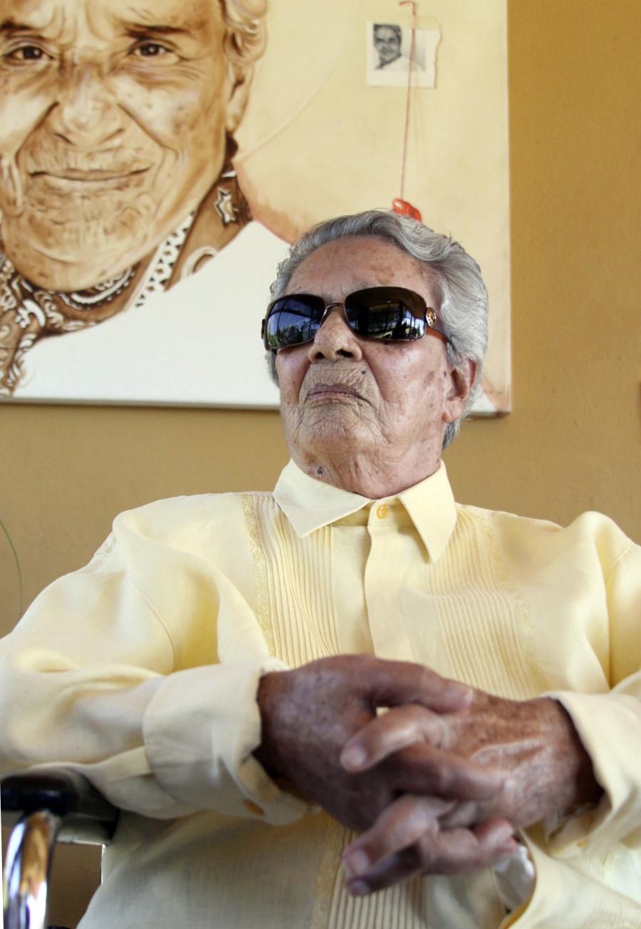 Chavela Vargas wears a yellow blouse and sunglasses and sits in front of a her painted portrait. 