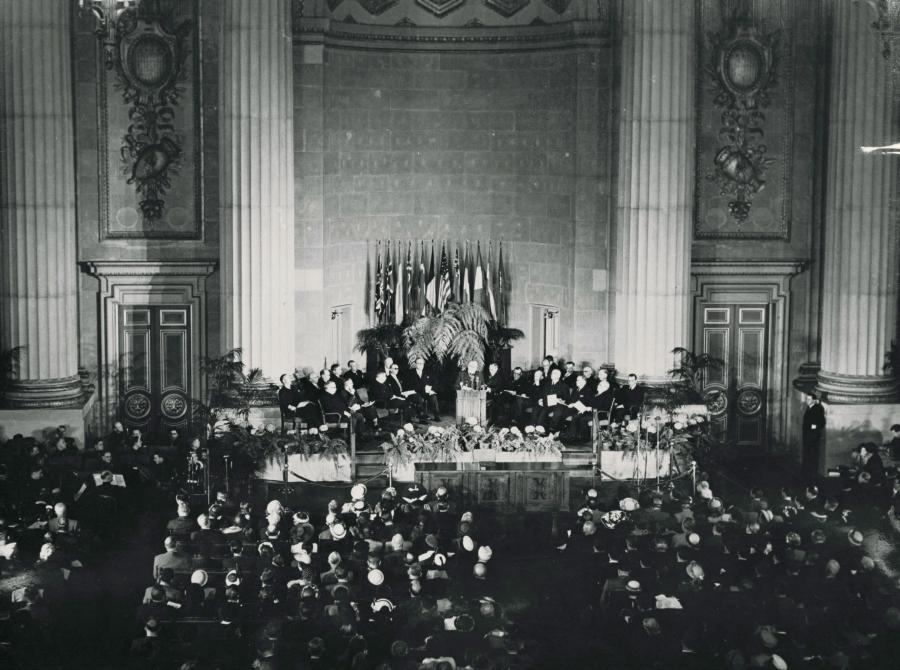 A historic photos shows dozens of delegates assembled in chairs around a podium, with more people seated around a lecturn. Behind there are giant columns. 