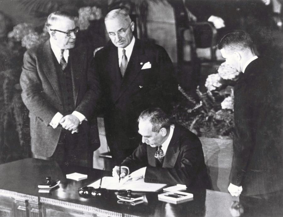 A man signs a piece of paper and two others look on in a historic black and white photo