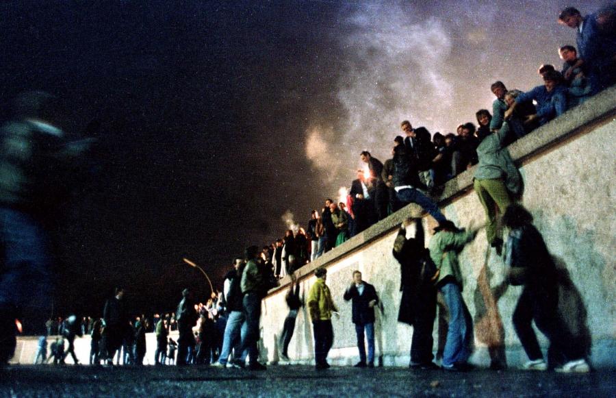 A crowd of people stand atop the Berlin Wall near Brandenburg gate in this 1989 photo. Behind them is smoke from what could be burning fires. 