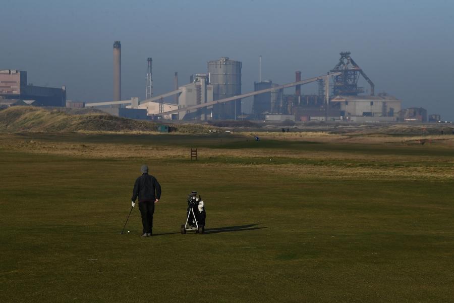 A man is show walking with a golf bag with an industrial steelworks plant in the background.