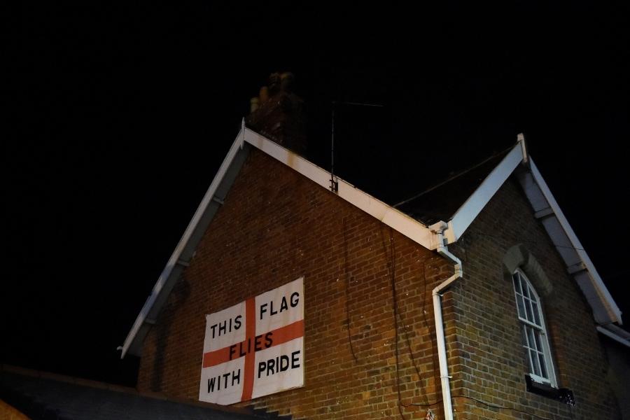 An England flag bearing the words: 'This flag flies with pride' is affixed to the side of a red brick house with white trim.