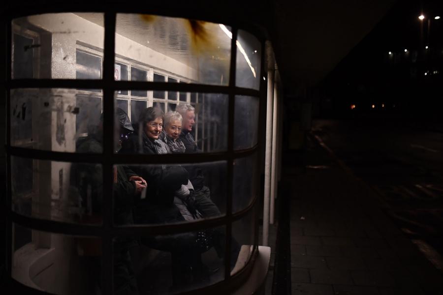 Several older people are shown leaning against the wall of a glass bus stop.