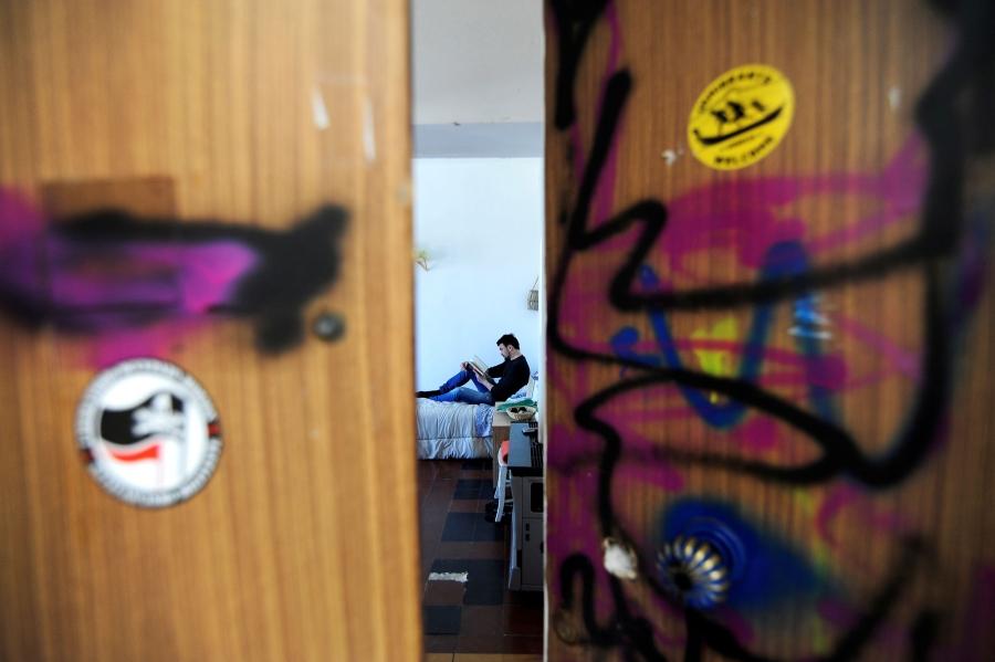 Alessandro Dus is shown through graffiti-painted doors, sitting on a bed reading a book.
