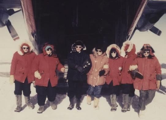 Six women wearing red parkas and a man in black pose for a photo.