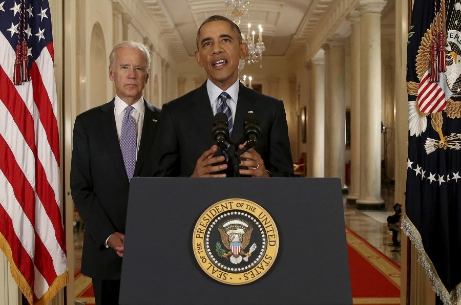 A black man stands behind a podium with the presidential seal. A white man stands behind him. There is an American flag in the corner.  