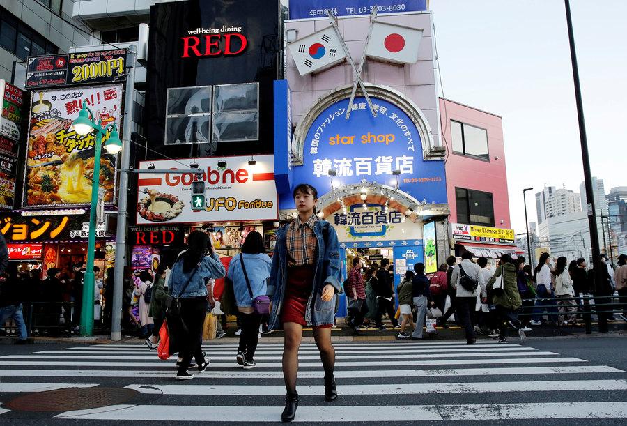 A woman is shown walking across a large street with billboard-clad buildings in the background.