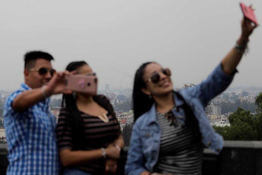 Blurry people in the foreground take a selfie with smog-covered city in the background.