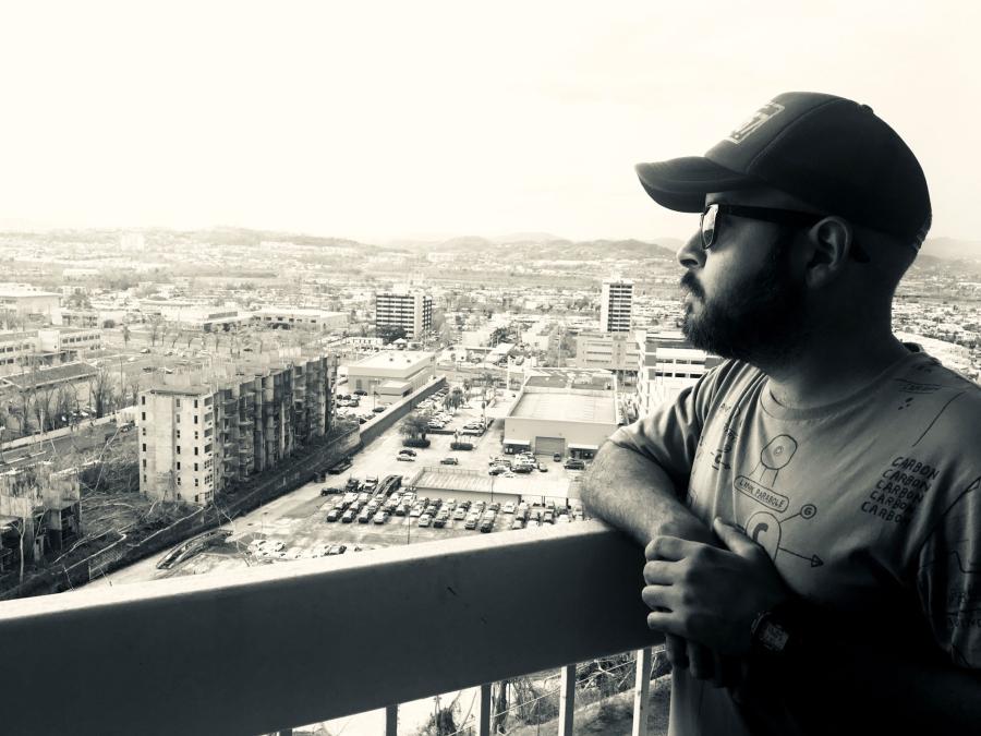 Gabriel Rodriguez leans on a balcony overlooking San Juan, Puerto Rico. HIs face is shadowed and he is wearing a baseball cap and sunglasses.