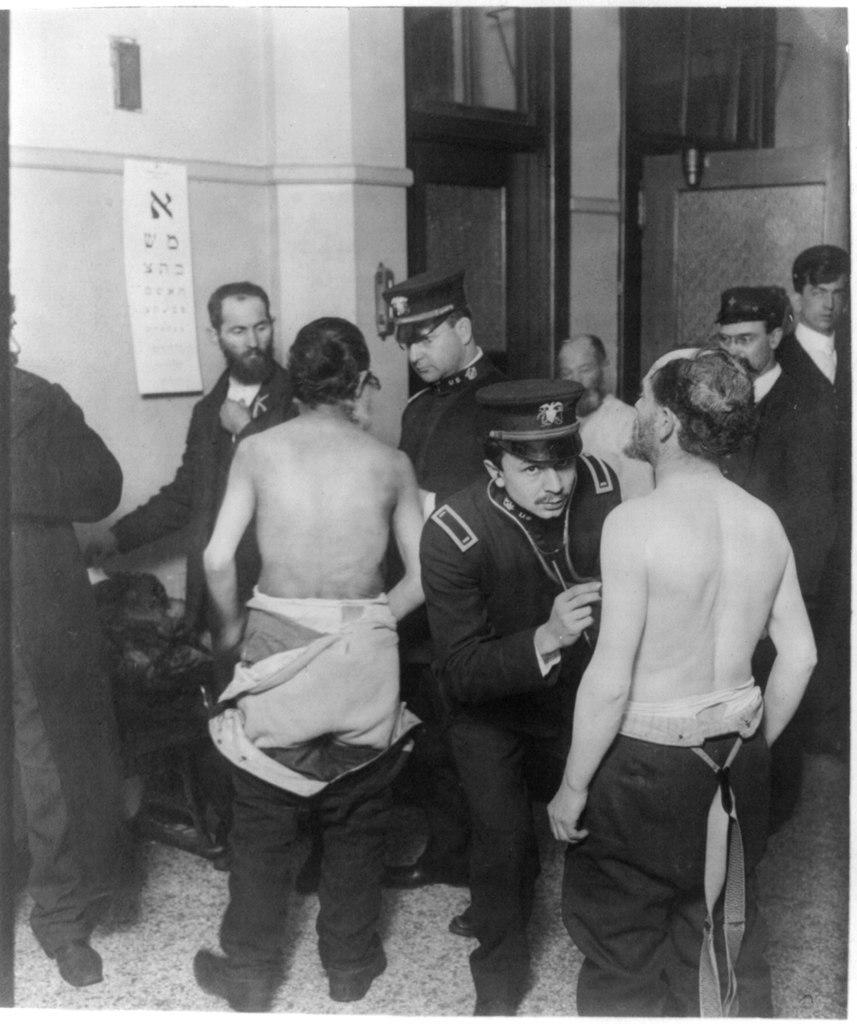 Photograph shows immigrants gathered in small room, two with shirts off being examined by physicians; eye chart written in Hebrew hangs on wall.