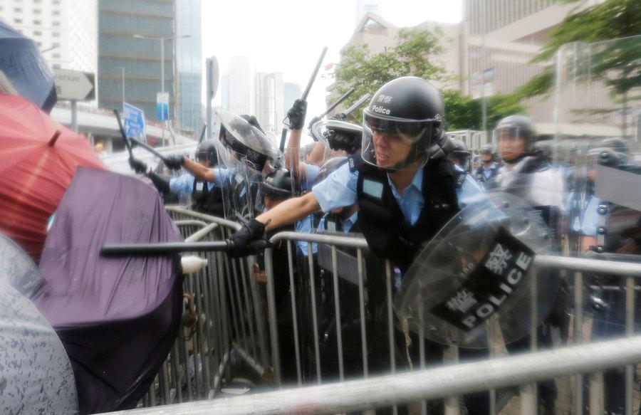 A police officer is shown with a helmet and clear face mask down, striking a person with an umbrella with his batton.