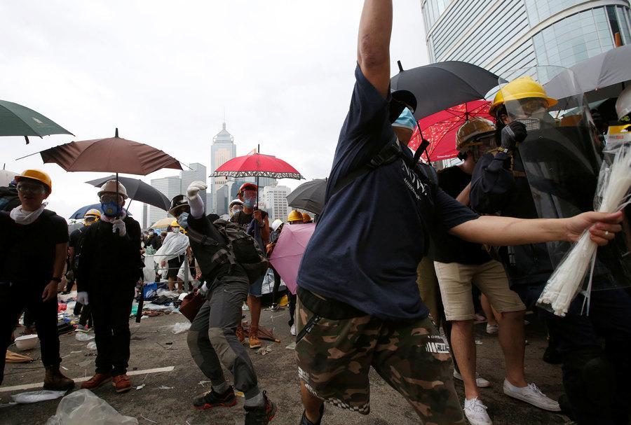 Several potesters are shown wearing masks and holding umbrellas with one person center frame who is about to throw and object.