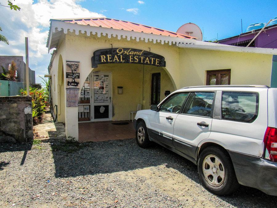 A Suburu car is parked outside of a yellow stucco building with a wooden sign that says "Island Real Estate on it."