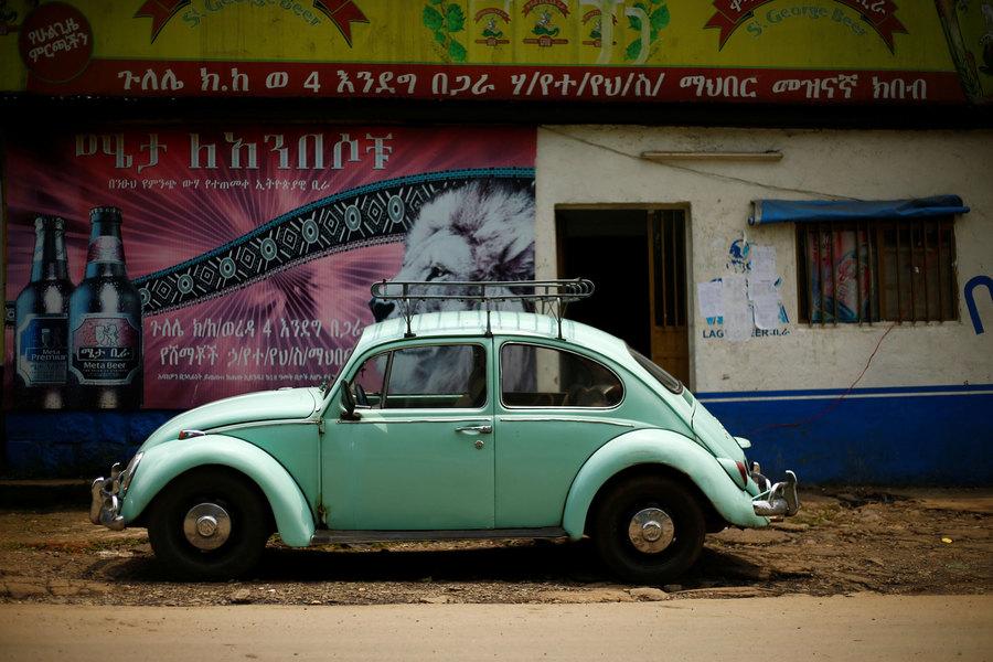 A greenish-blue colored Volkswagen Beetle is shown with a metal rack on top and parked in front of a store.