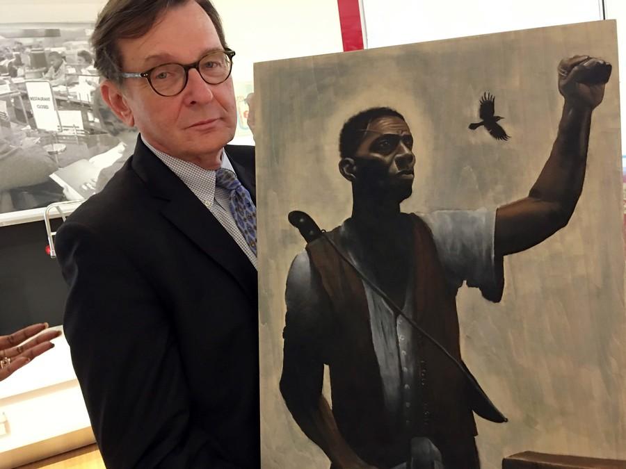 A white man in glasses and a suit holds a painting of a black man with a raised fist wearing a shirt and vest. 