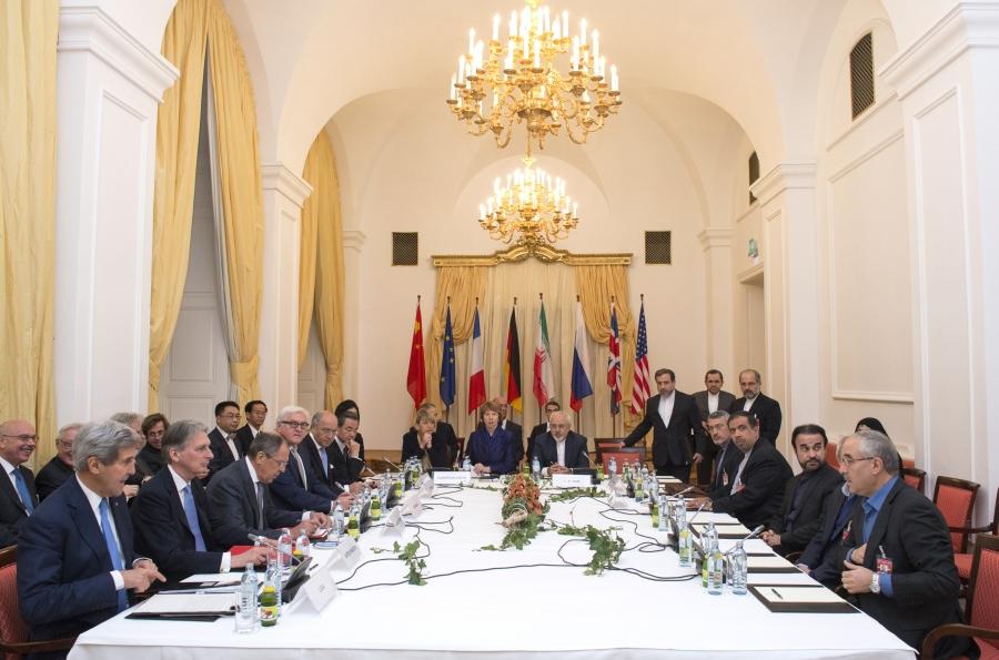 Men and women sit around a negotiating table with flags in the background. 