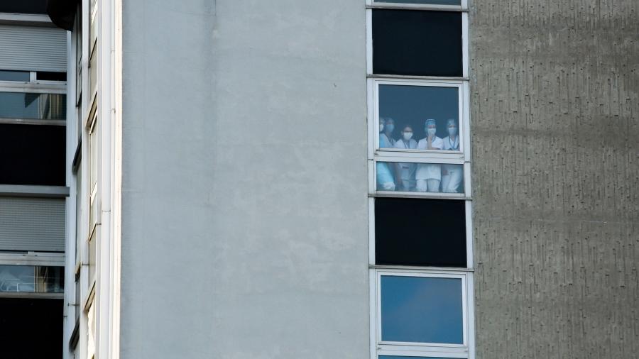 Medical personnel are shown standing in a window wearing protective medical gear and photographed from far outside of the building from below.