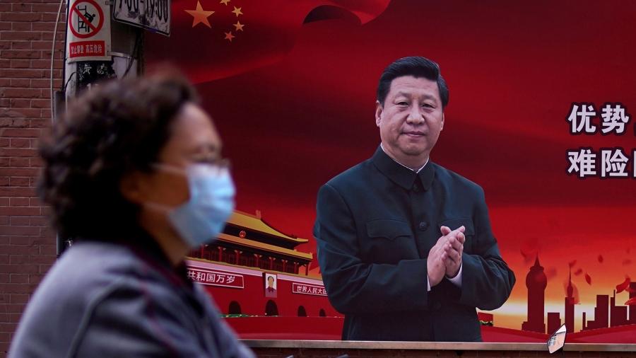 A woman is shown in soft focus and wearing a protective mask while walking past a portrait of Chinese President Xi Jinping on a red background. 
