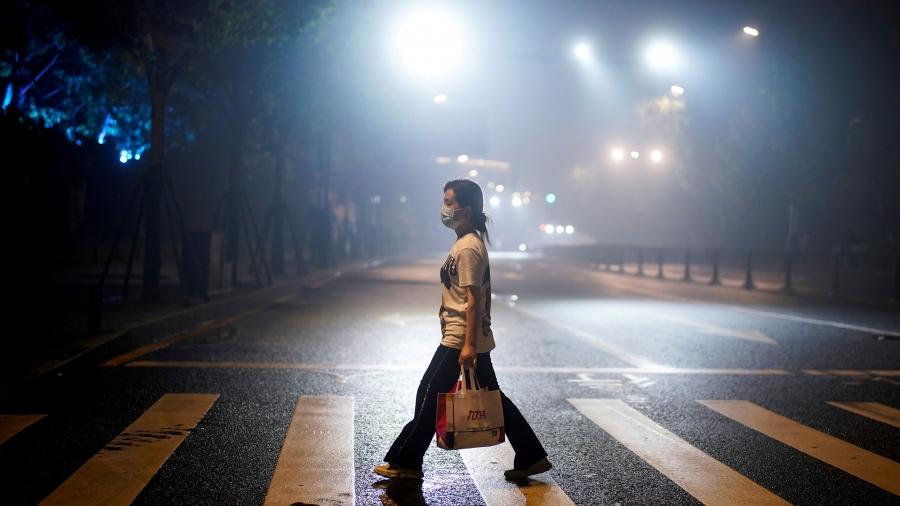 A woman wearing is shown walks on a street in the crosswalk of a street while carrying a bag and wearing a protective face mask with street lights shining above.
