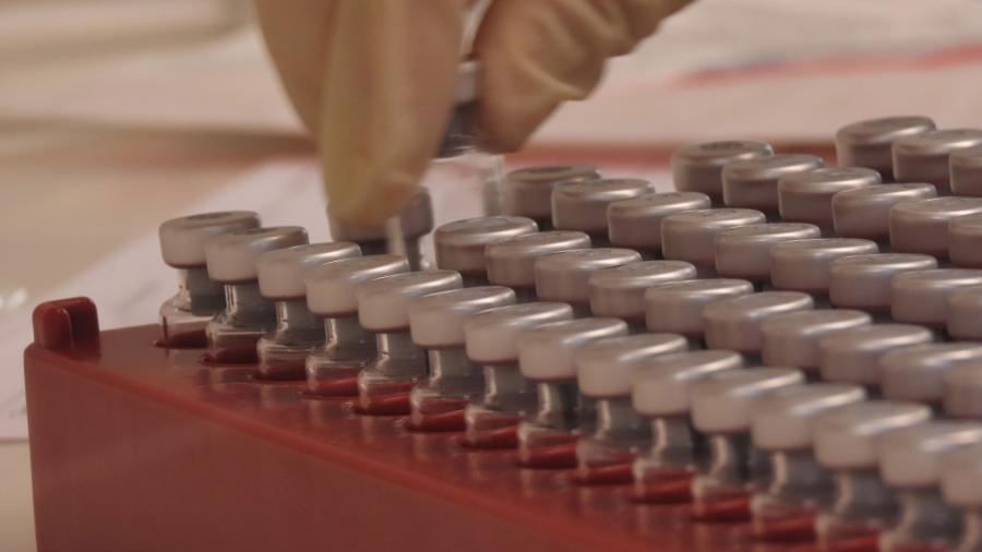 A red tray of dozens of vials is shown with a hand picking one of the vials up.