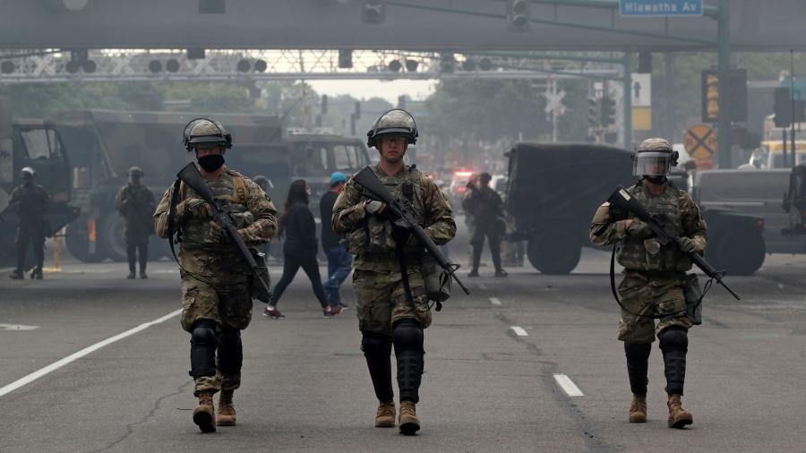 Several US National Guard members are shown wearing military fatigues and carrying weapons while walking in a street.