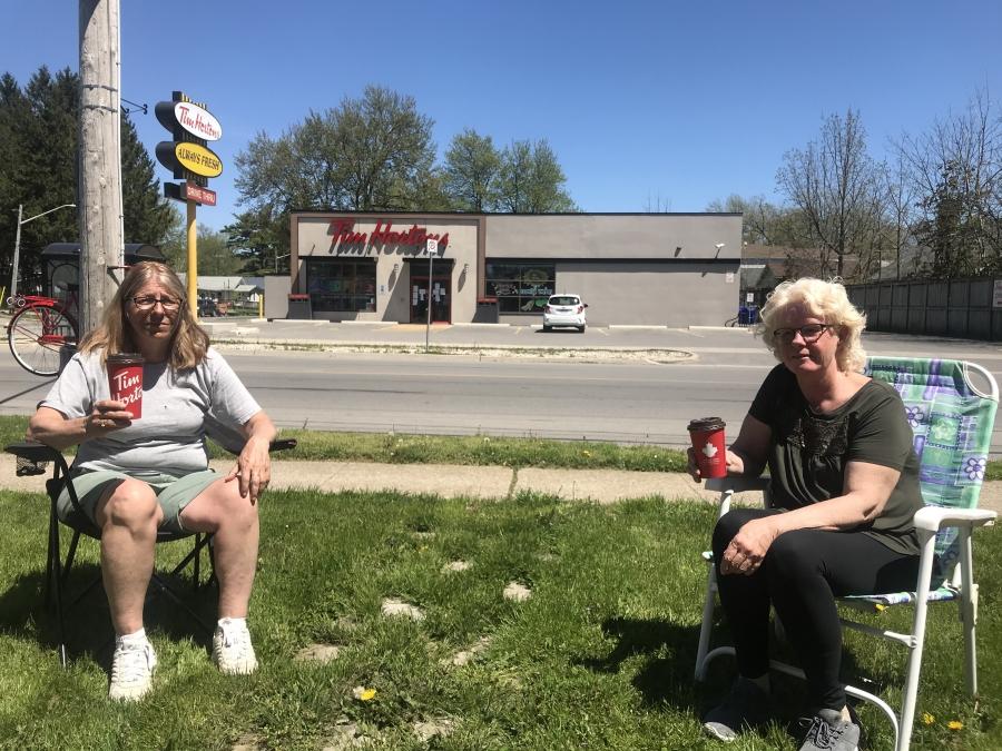 Two women sit in lawn chairs and drink a coffee near a coffee shop. 