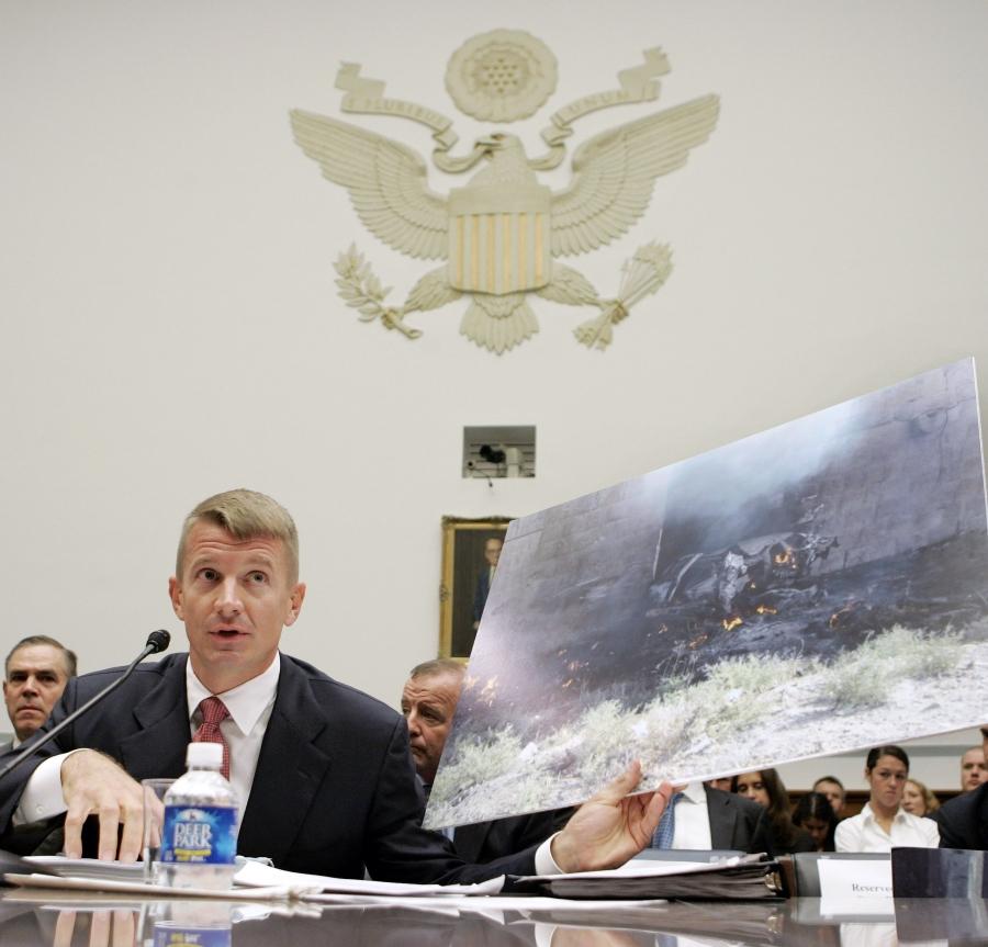 A man sits at a desk in front of an emblem of the United States, holding a large photograph