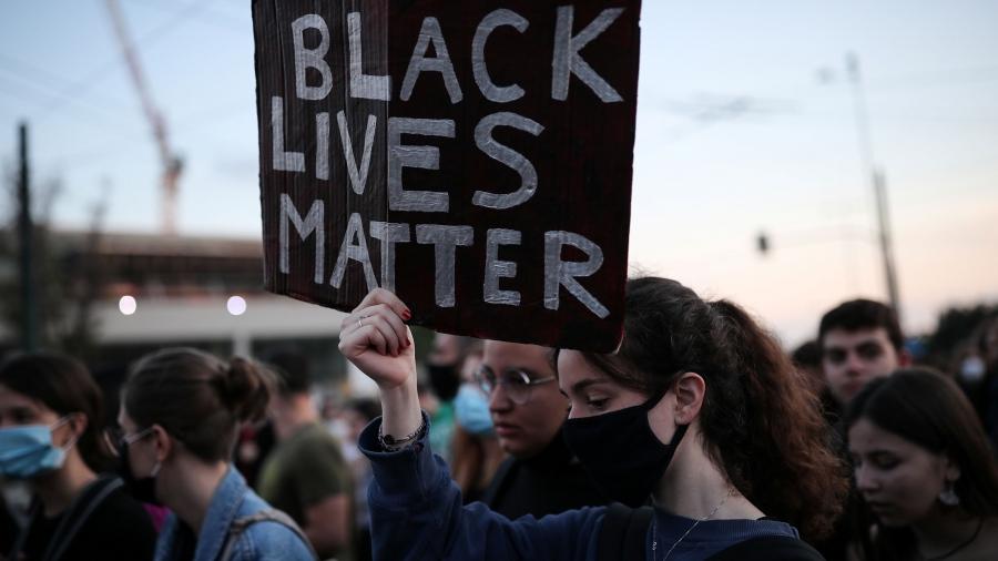 A large group of people are shown in a crowd with one woman in the center carrying a sign with "Black Lives Matter" written on it.
