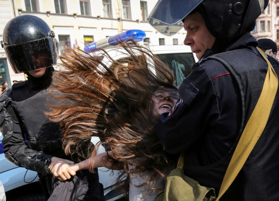 Two Russian police officers are shown with helmets and dark visors holding a woman who is resisting.