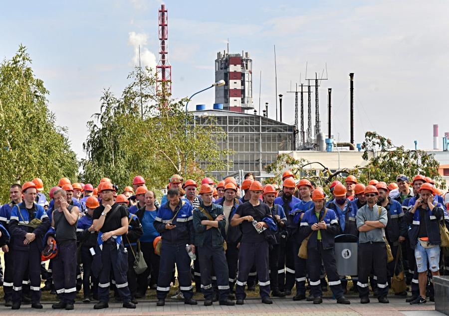A large group of men with hard hats standing outside of their work's manufacturing facility. 