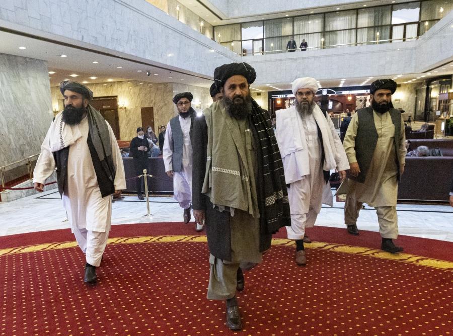 A group of men wearing Afghan traditional clothes walk across a reddish carpet in a lobby.