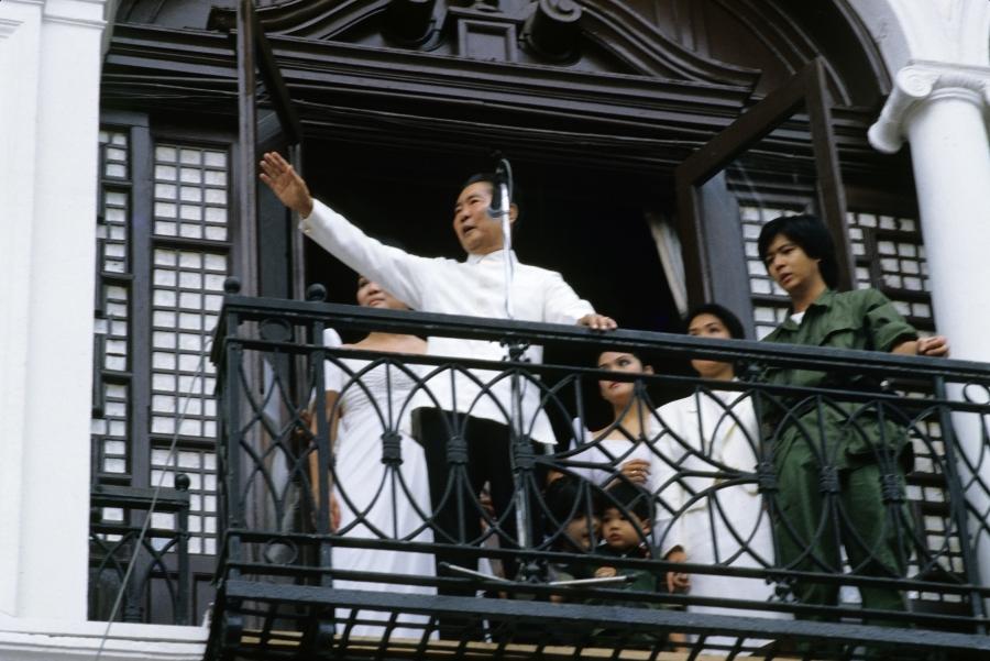 Ferdinand Marcos, dressed in white traditional Philippine shirt, raises his hand and speaks into a microphone to supporters; beside him in a green jumpsuit is his son, Bongbong Marcos.