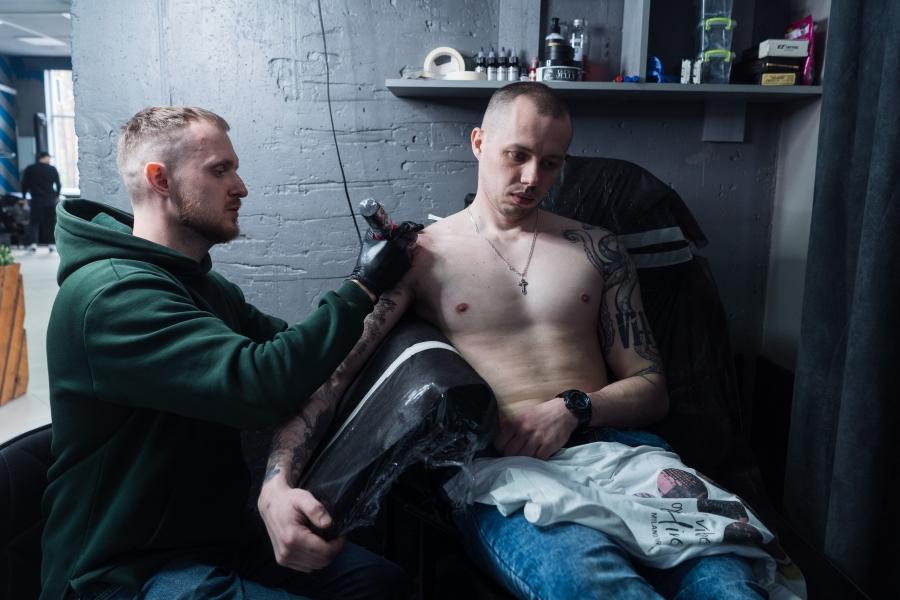 A man wearing a green hoodie while tattooing a shirtless man in a chair