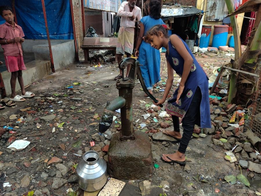 A young girl utilizes a water pump in the middle of the street in a small neighborhood.