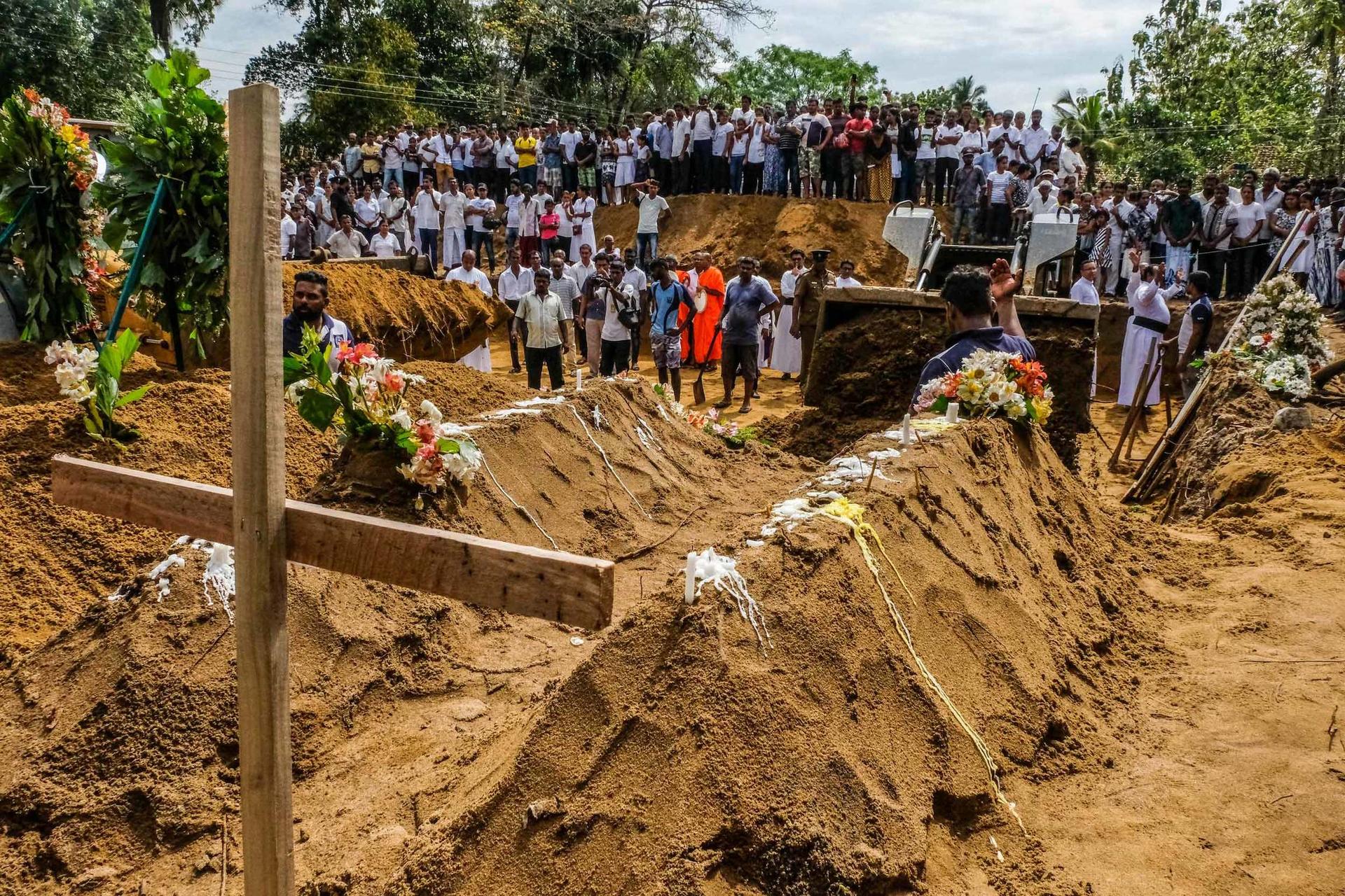 A crowd of people watch as a backhoe moves dirt over caskets during a mass funeral