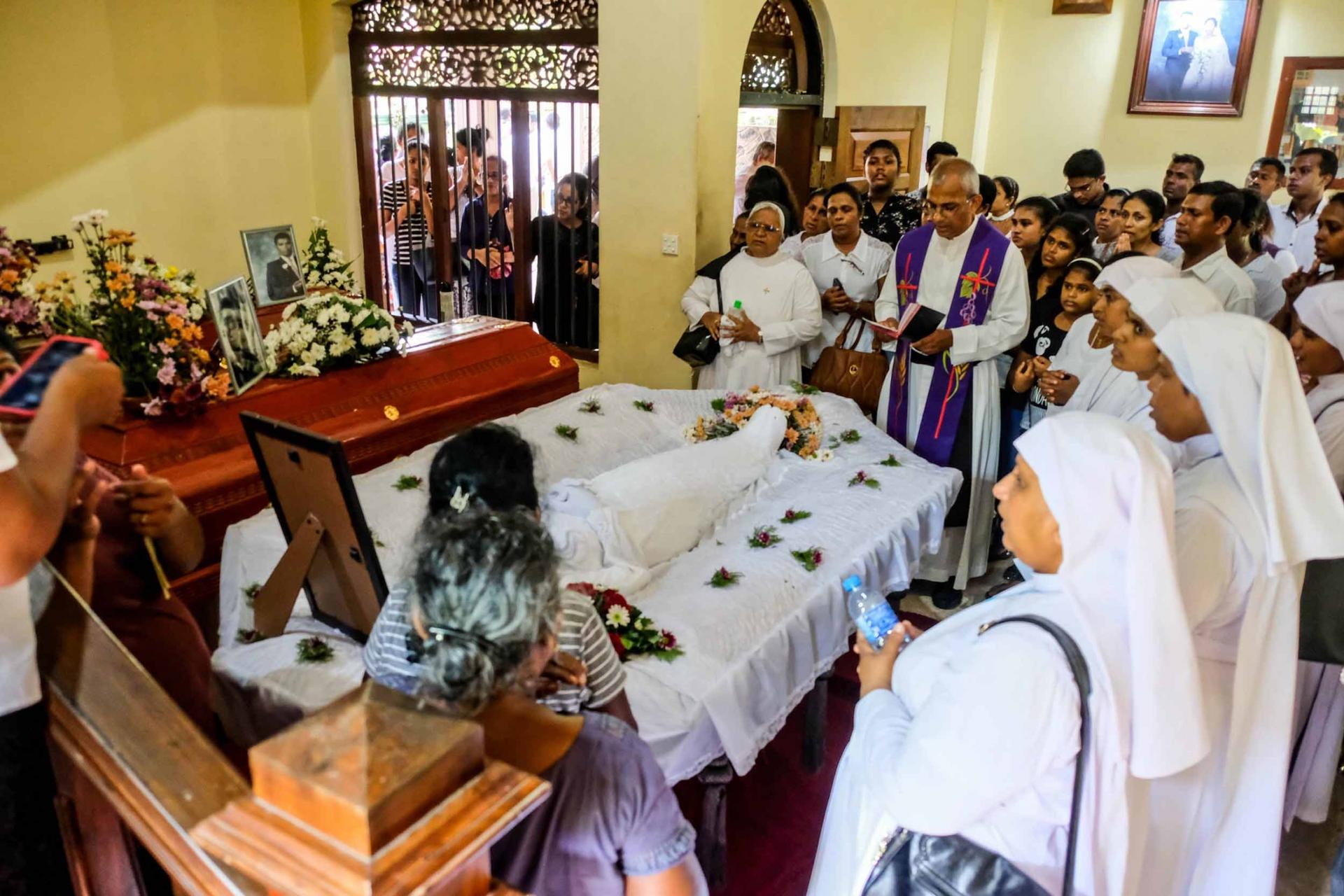 Family and friends gather around a casket in a small home while a priest reads a service