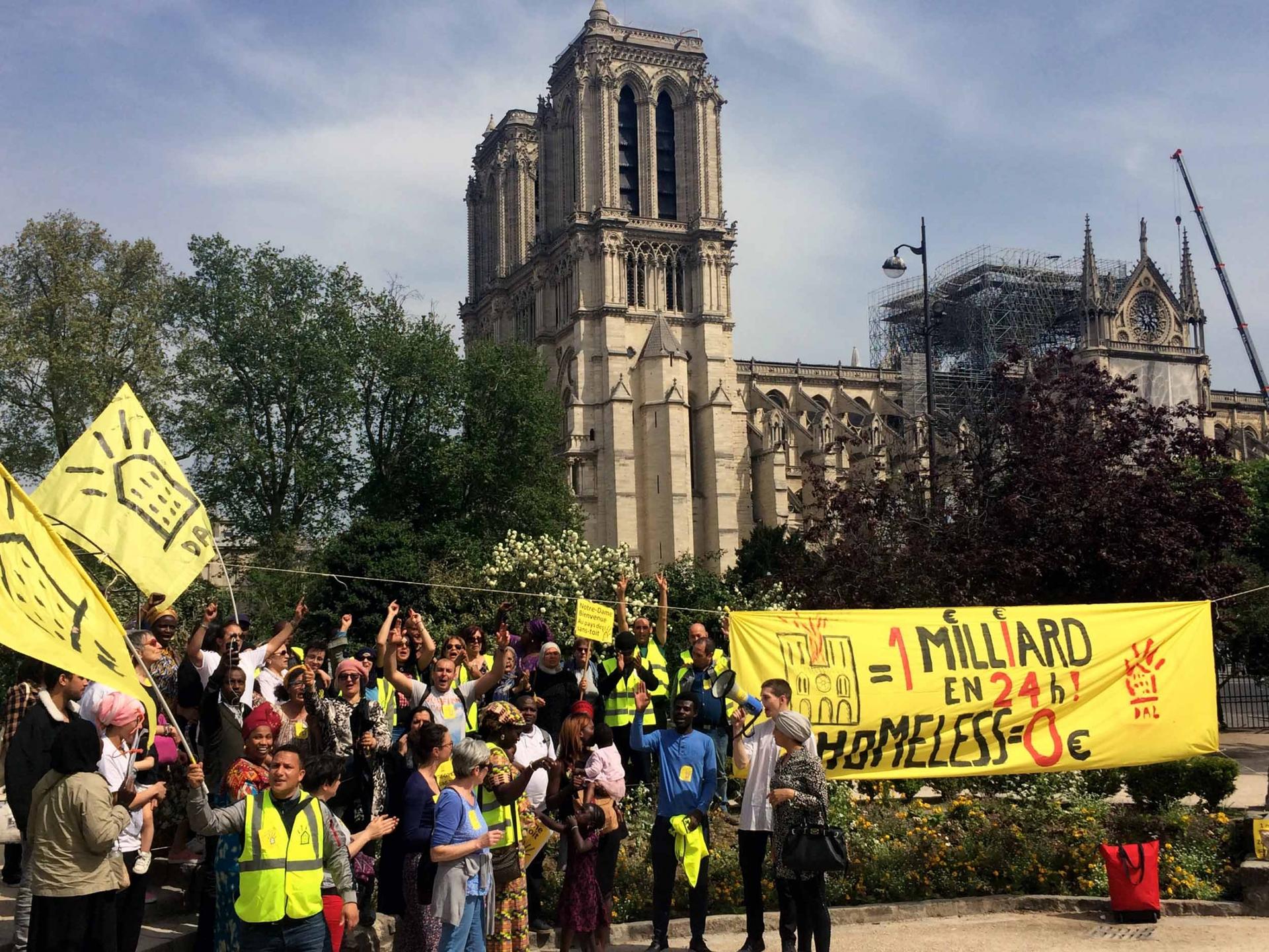 A crowd of people hold yellow signs and a banner. The towers of Notre-Dame are visible in the background. 