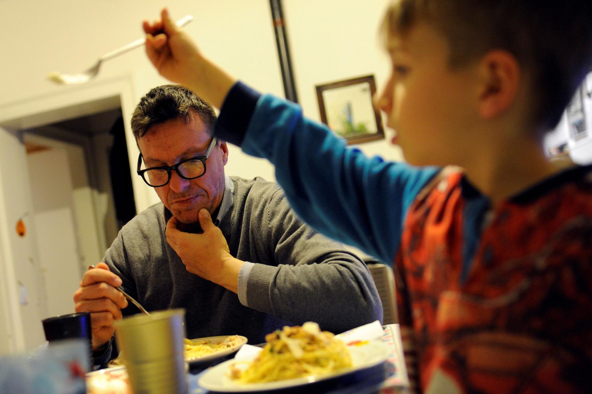 A young boy is shown in the nearground of the photo holding up a fork with his father sitting in the background eating from a plate of food.