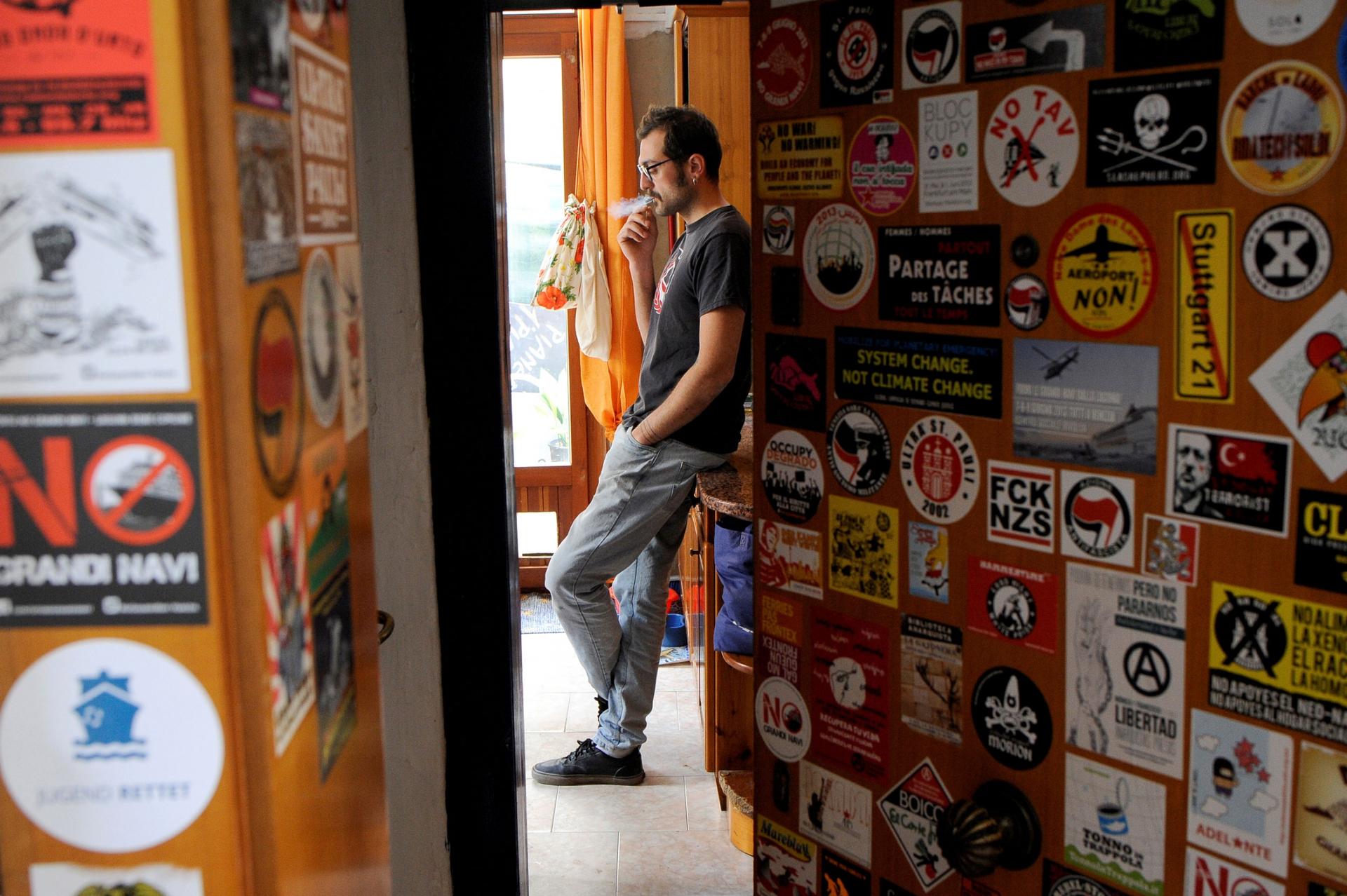 A man is seen through a sticker-covered doorway, leaning up against a dresser and smoking a cigarette.