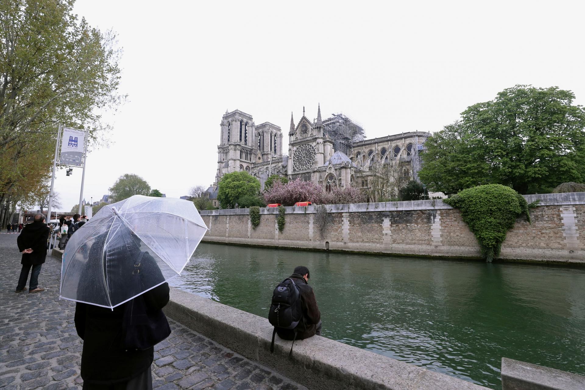 A person holding a clear umbrella stands across the river looking at a burned Notre-Dame Cathedral.