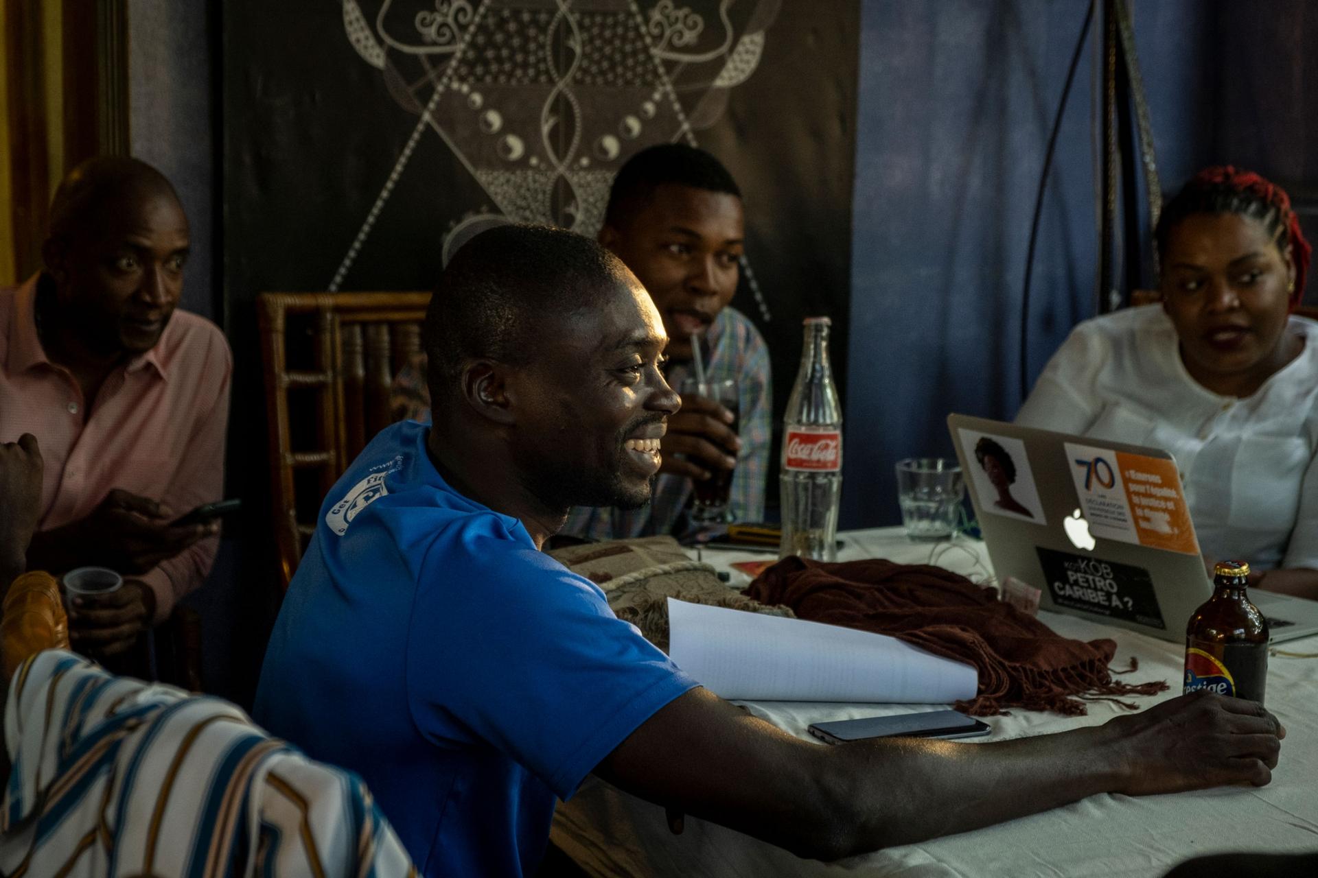 A group of young Haitians are shown sitting around a table with computers, Coke and beer bottles on top.